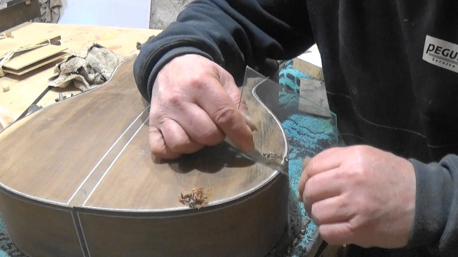 Mãos de luthier raspando binding com raspilha de aço em movimento de fora para dentro ao longo da borda do violão