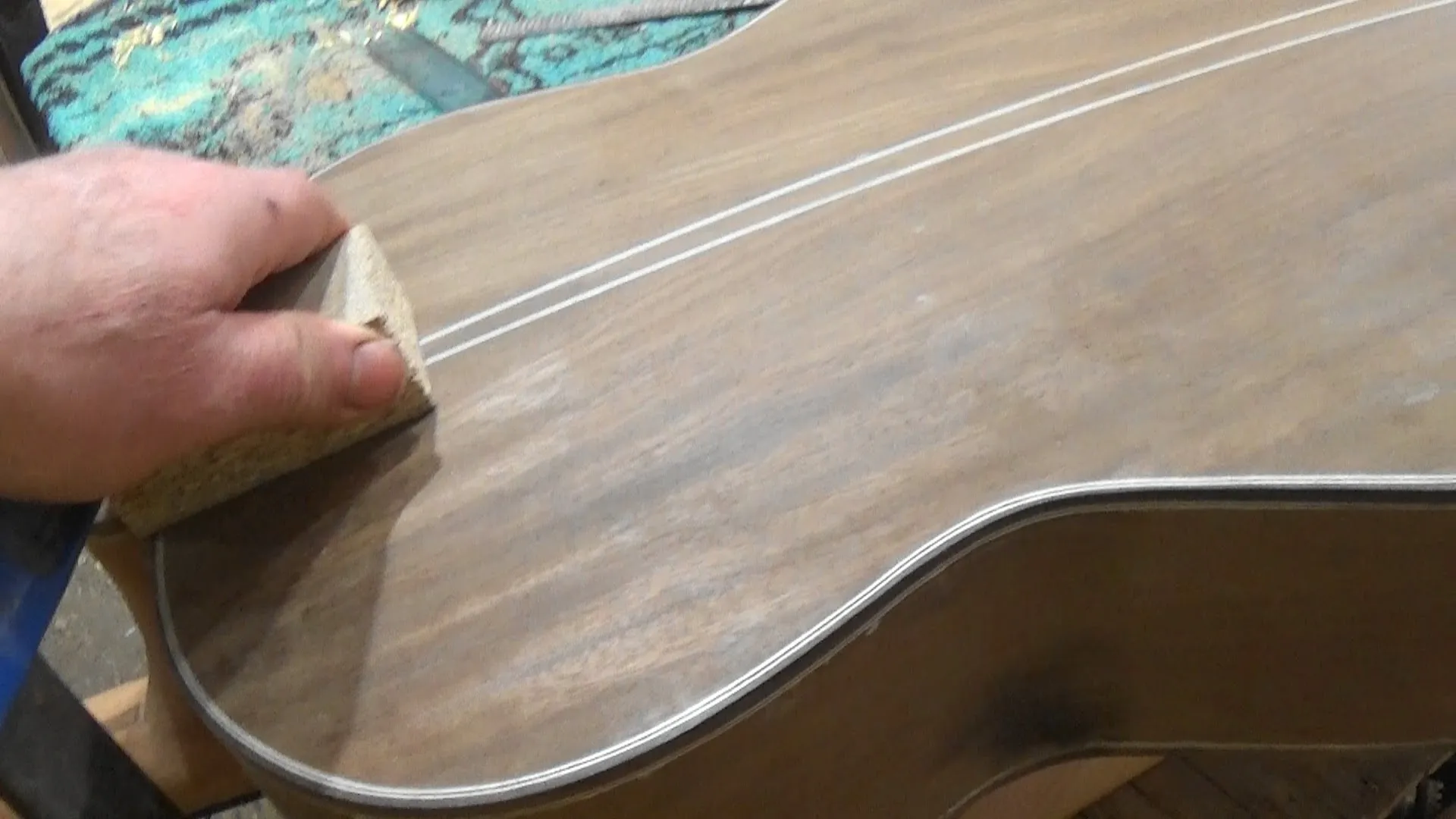 Luthier lixando o tampo do violão com taco de madeira seguindo sequência progressiva de grãos antes do acabamento